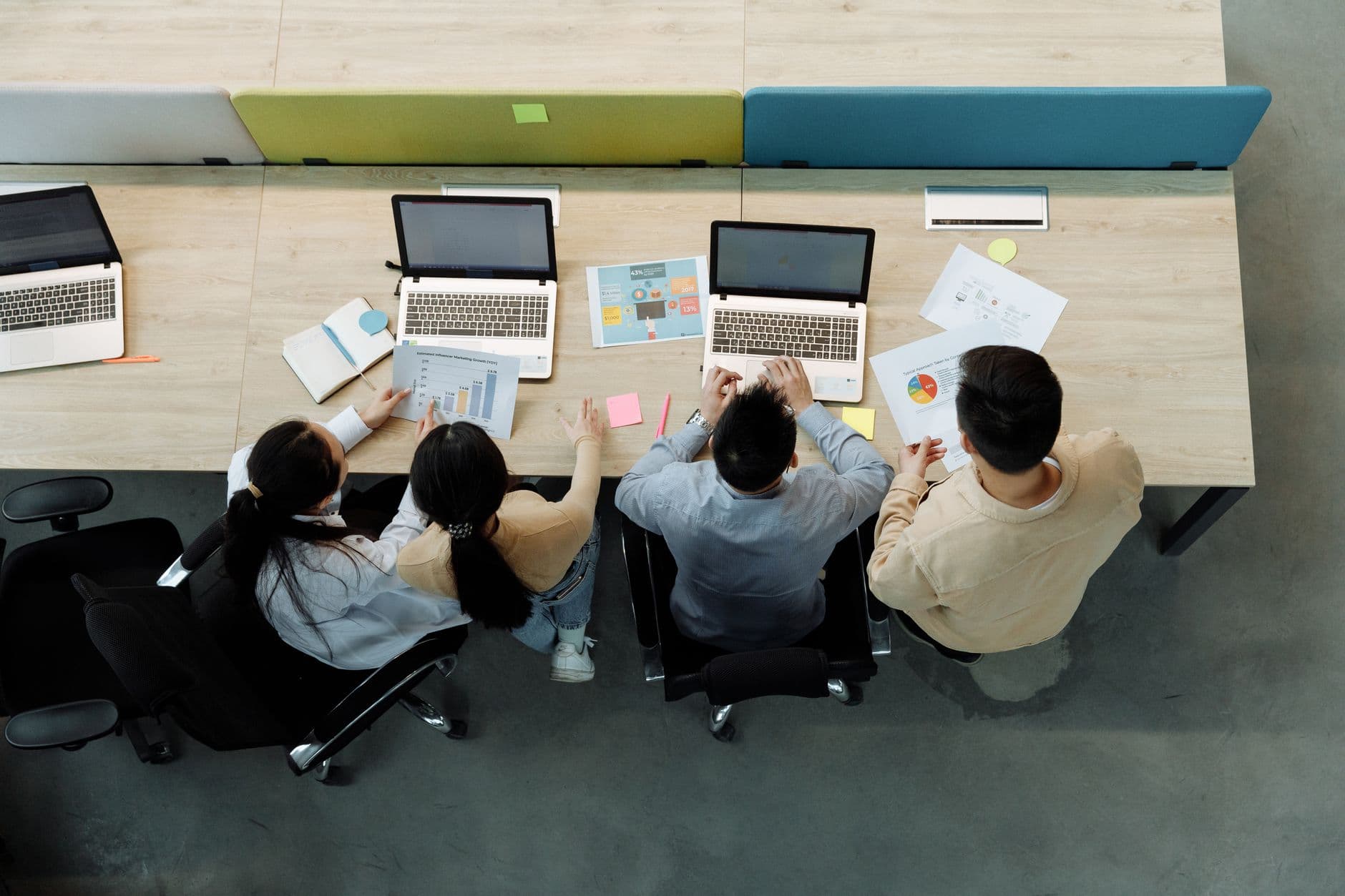 Four colleagues working together at a shared office desk with laptops and charts